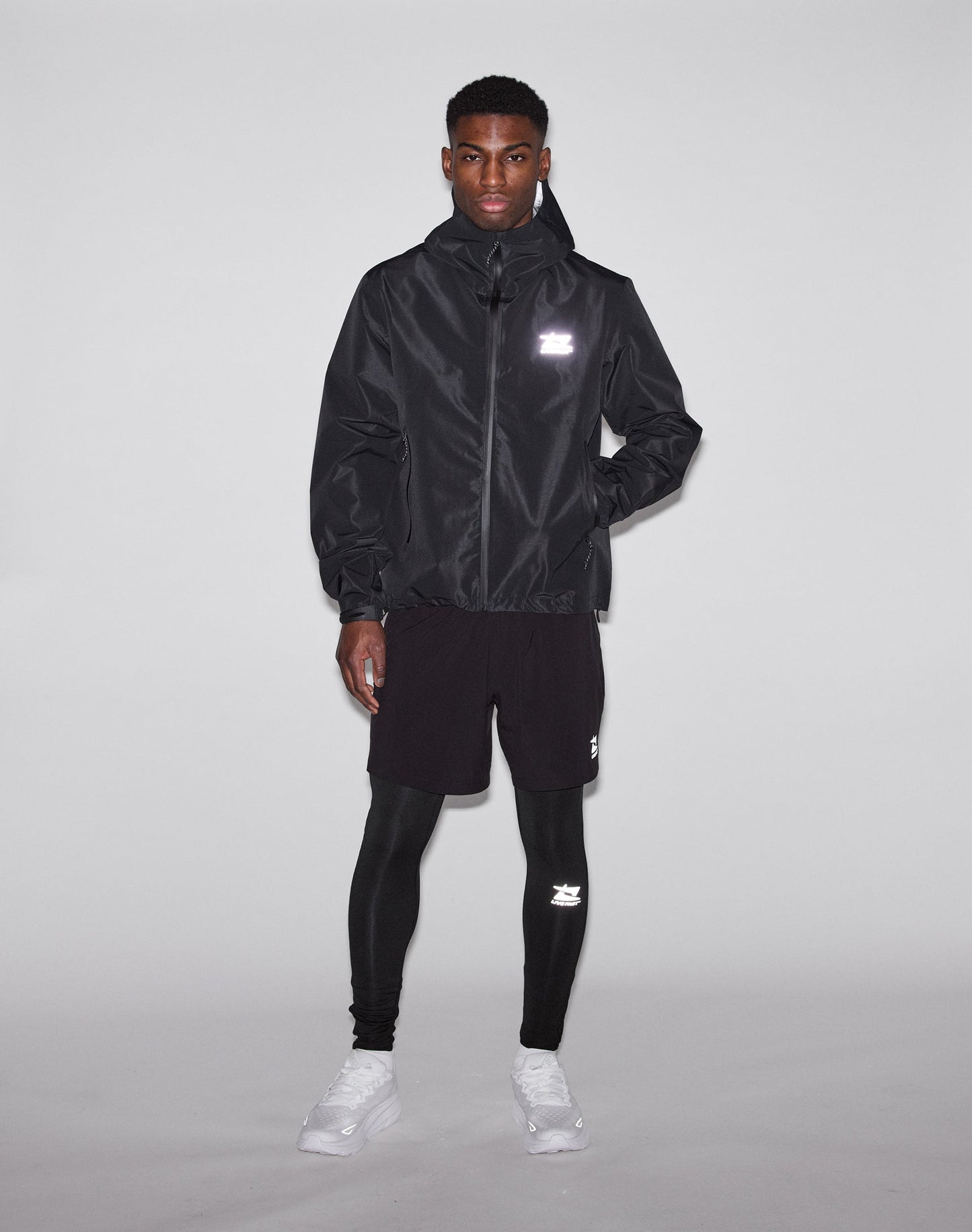 A man stands against a plain background wearing the LIVE FAST DIE YOUNG Live Fast Performance Rain Jacket with wasserdichte Nähte, black shorts and leggings, white sneakers, and reflective logos. His hands are at his sides.