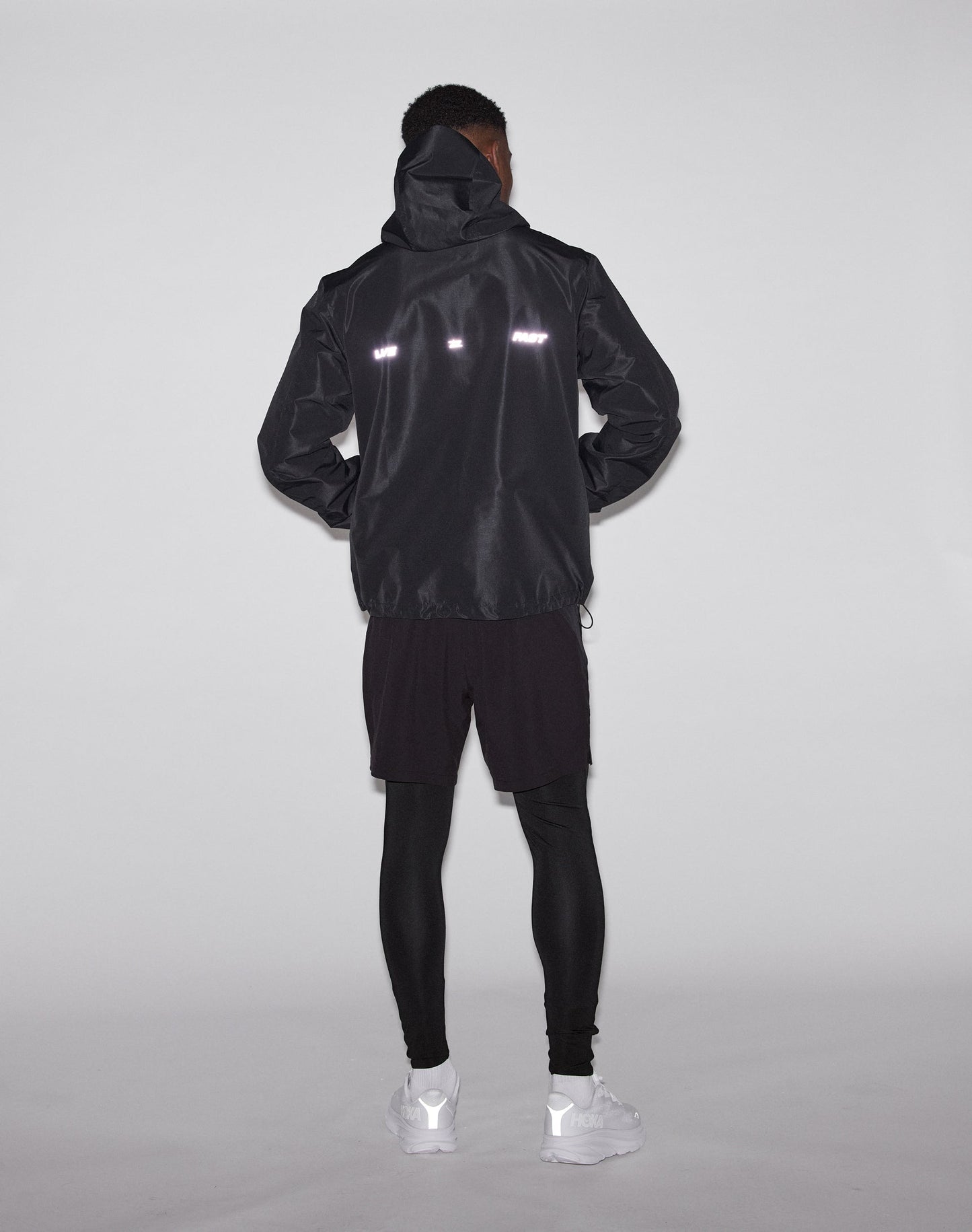 A person stands facing away, wearing the LIVE FAST DIE YOUNG Live Fast Performance Rain Jacket with waterproof seams and reflective strips, black shorts over leggings, and white sneakers against a plain light background.