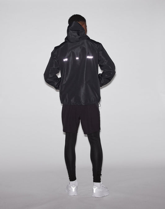 A person stands facing away, wearing the LIVE FAST DIE YOUNG Live Fast Performance Rain Jacket with waterproof seams and reflective strips, black shorts over leggings, and white sneakers against a plain light background.