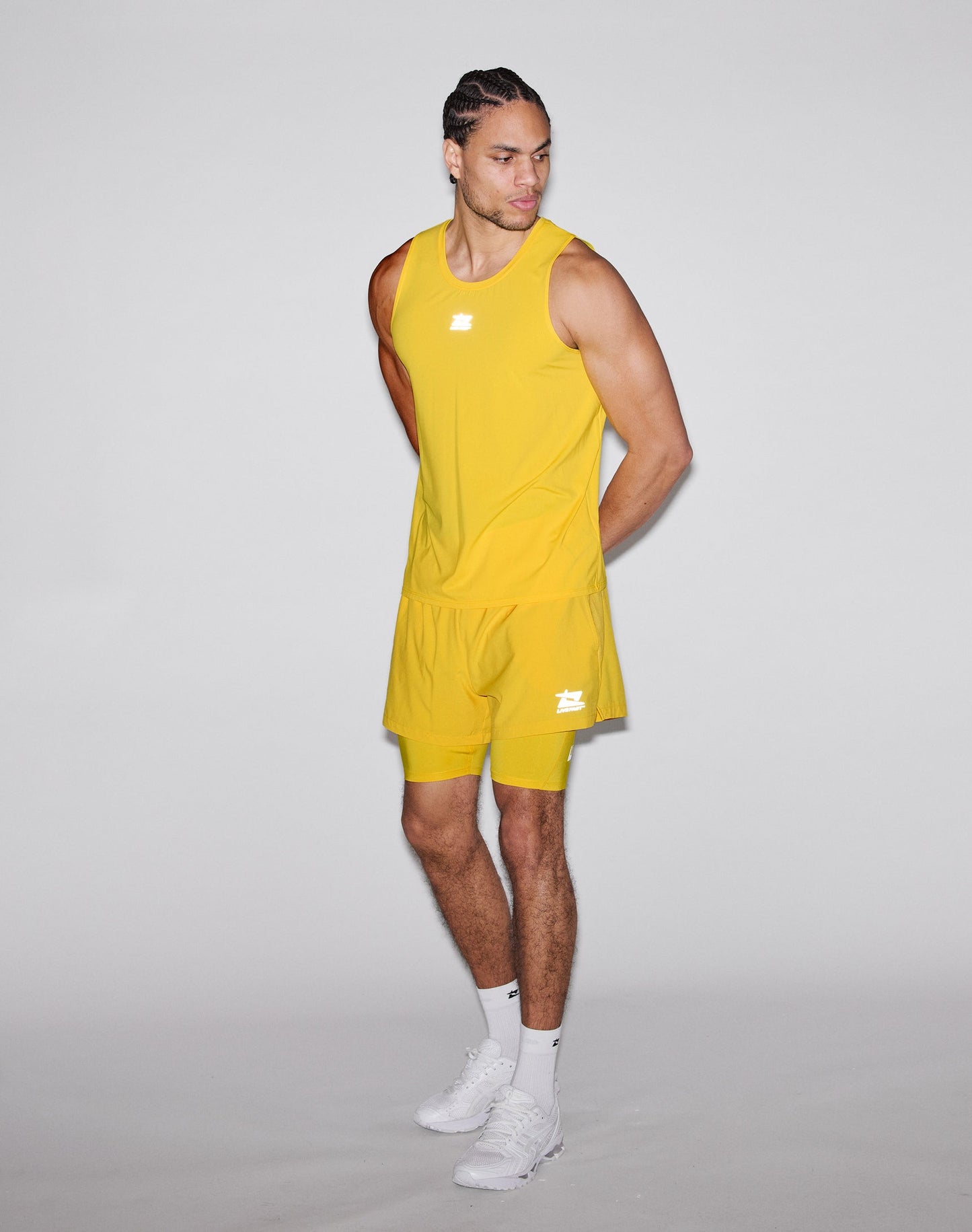 A man models the LIVE FAST DIE YOUNG Live Fast Performance Tank in bright yellow with matching shorts featuring reflective print, standing against a plain light gray background and looking left with his hands behind his back.