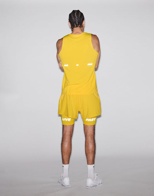 A person with braided hair wears the LIVE FAST DIE YOUNG Live Fast Performance Tank in yellow and matching shorts with reflective LIVE FAST print, plus white socks and sneakers. They face away from the camera with arms crossed against a plain background.