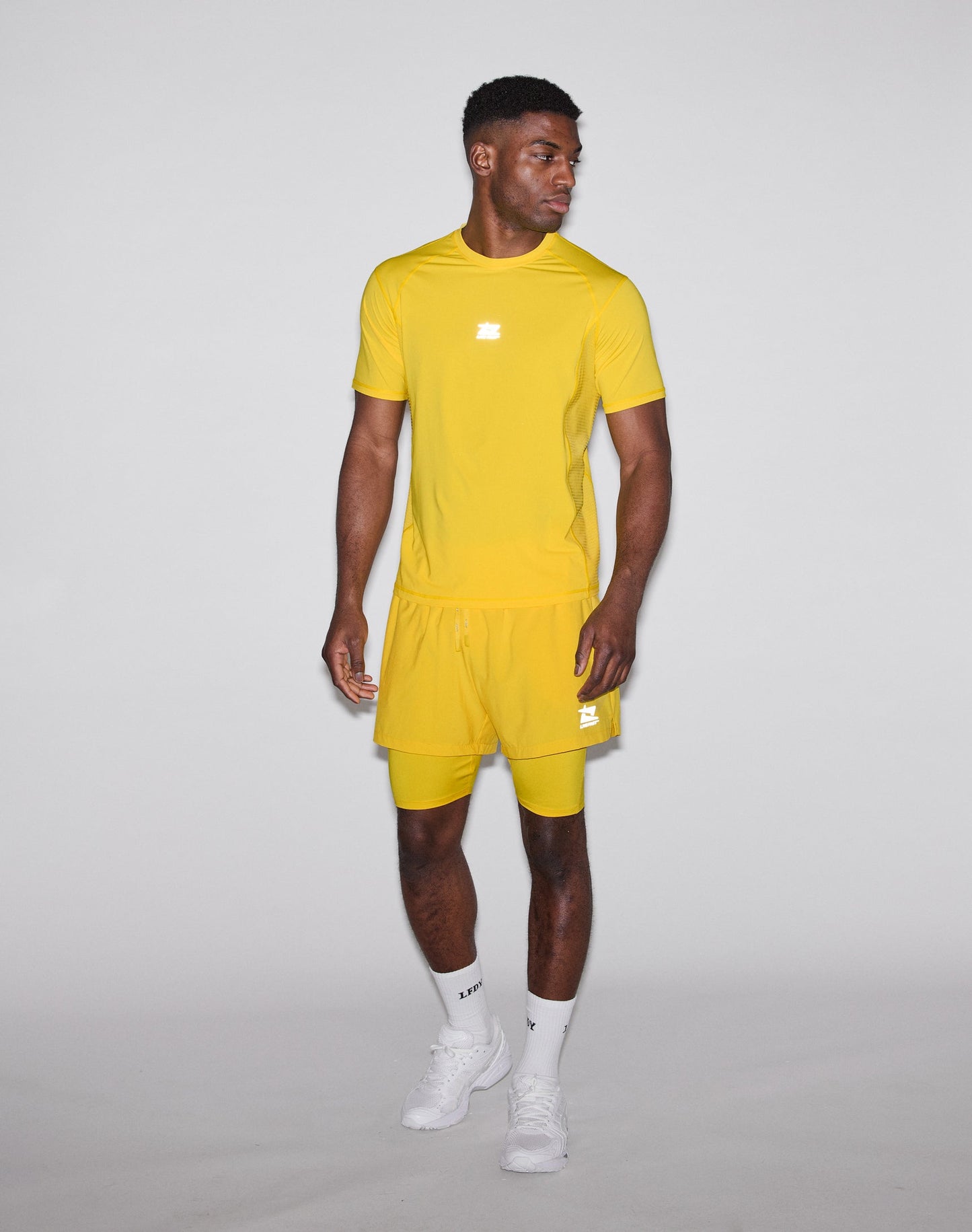 A man stands against a plain light background wearing the LIV FAST DIE YOUNG Live Fast Performance Tee in bright yellow with matching shorts, both made from comfortable Nylon Spandex. He looks to his left with one hand by his side, paired with white socks and sneakers.
