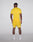 A man faces away from the camera in a slim-fit, bright yellow Live Fast Performance Tee by LIVE FAST DIE YOUNG, paired with matching shorts featuring LIVE and FAST, plus white socks and shoes. Reflective print details pop against a plain background.