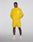 A man stands against a plain background wearing the bright yellow Live Fast Performance Running Jacket by LIVE FAST DIE YOUNG with reflektierender Frontprint, matching shorts, white socks, and white sneakers. Hands in pockets, he looks at the camera.