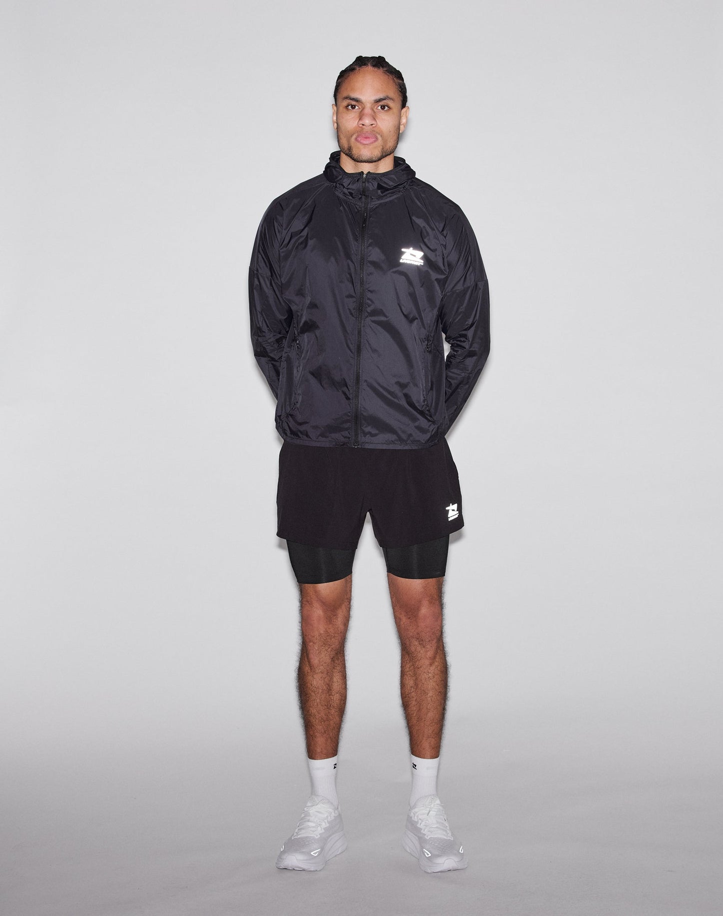 A man stands facing forward against a plain light background, wearing a LIVE FAST DIE YOUNG Live Fast Performance Running Jacket with Kapuze mit Visor, black shorts over black leggings, and white athletic shoes. His hands are behind his back.