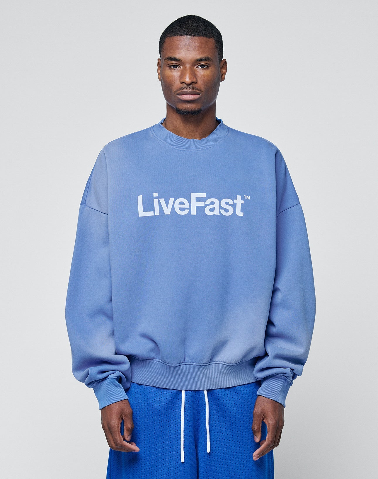 A man faces forward against a plain light gray background, wearing the Basic LiveFast Sweater from LIVE FAST DIE YOUNG in blue with “LiveFast” white text, paired with blue athletic shorts featuring white drawstrings.