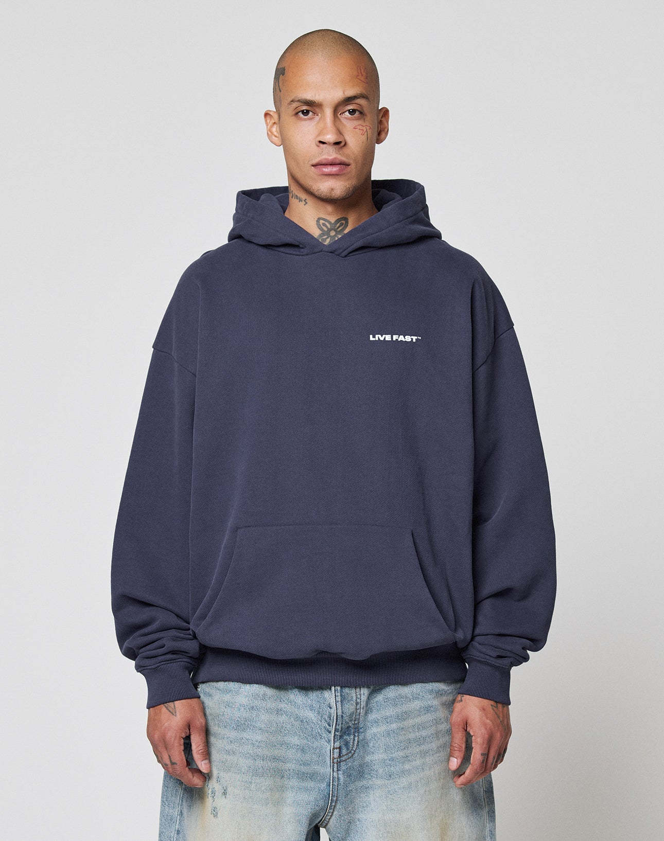 A person with a shaved head wears the Live Fast Plain Hooded by LIVE FAST DIE YOUNG in dark blue, featuring an oversize fit and front print, paired with light blue jeans, standing against a plain light background.