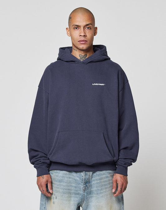 A person with a shaved head wears the Live Fast Plain Hooded by LIVE FAST DIE YOUNG in dark blue, featuring an oversize fit and front print, paired with light blue jeans, standing against a plain light background.