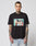 A man with neck and arm tattoos wears the LIVE FAST DIE YOUNG LFDY Sixers Tee, a regular fit black cotton T-shirt with a vibrant two-people-at-a-table graphic. He pairs it with light blue jeans against a white background. Care instructions on label.