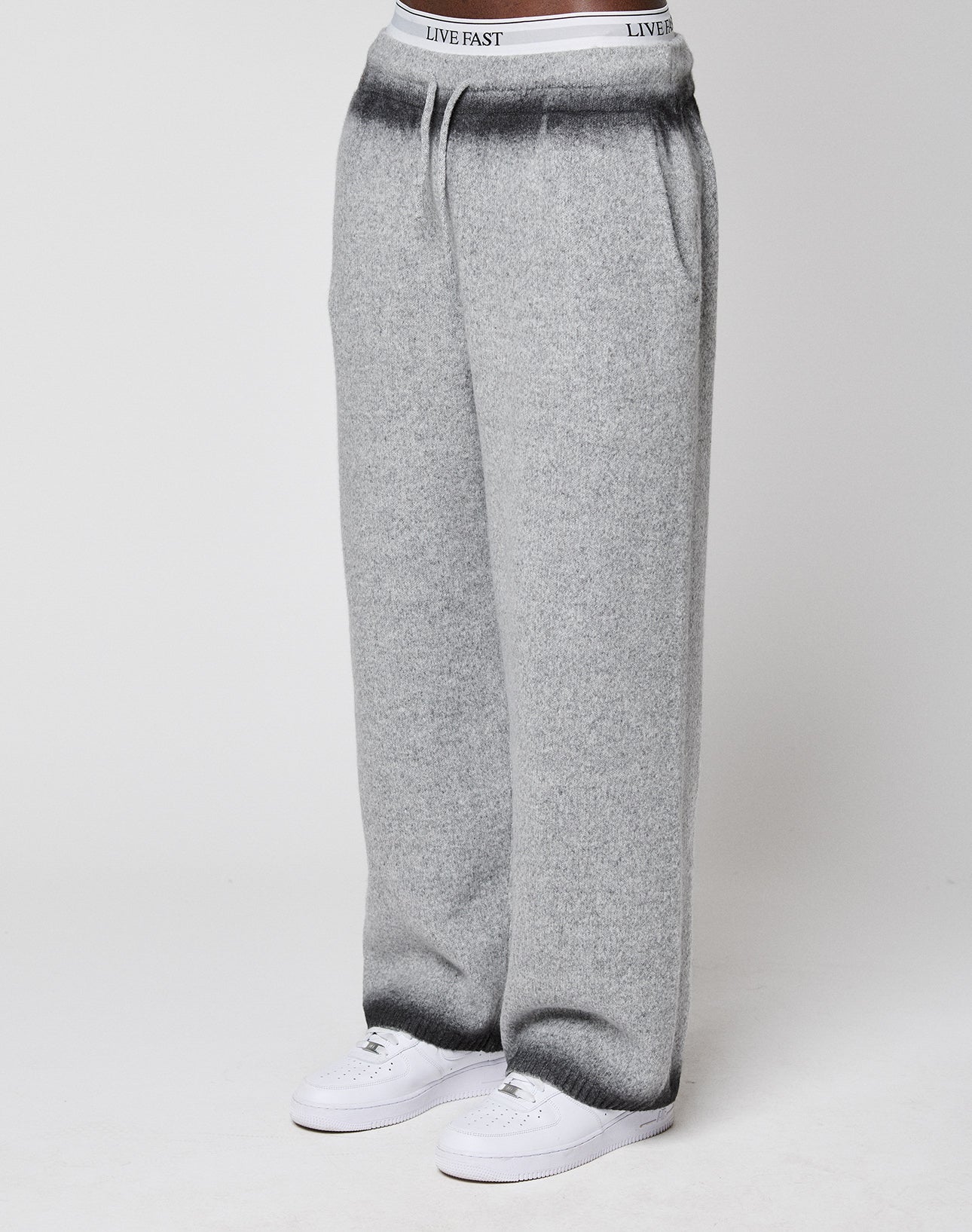 Someone wears LIVE FAST DIE YOUNG's Live Fast Boucle Pants—wide fit, light gray, straight leg with elastic waistband labeled "LIVE FAST"—with white sneakers, standing against a plain background.