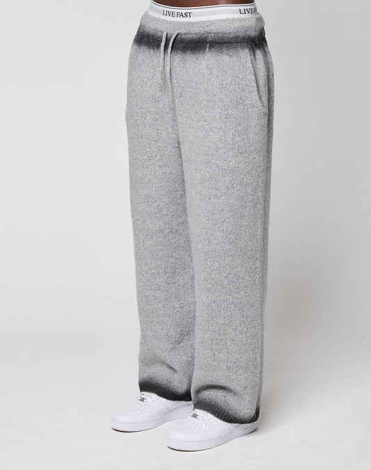 Someone wears LIVE FAST DIE YOUNG's Live Fast Boucle Pants—wide fit, light gray, straight leg with elastic waistband labeled "LIVE FAST"—with white sneakers, standing against a plain background.