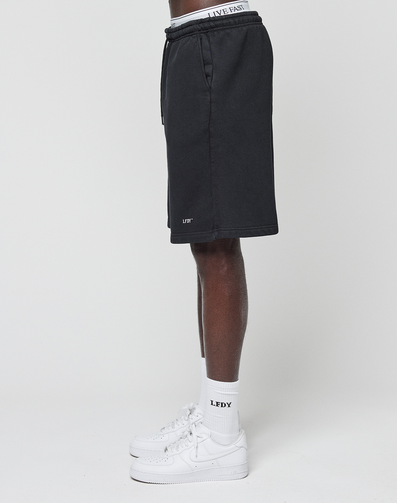 A person stands sideways against a plain white background wearing LIVE FAST DIE YOUNG Basic Shorts with a logo print near the hem and an elastic waistband that says LIVE FAST, paired with white sneakers and LFDY socks.