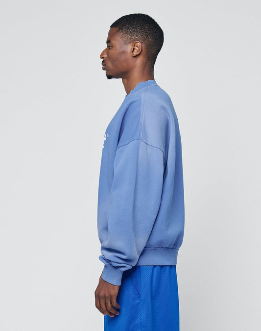A man stands in profile against a plain light background, wearing the LIVE FAST DIE YOUNG Basic LiveFast Sweater and matching blue shorts with a subtle handgefertigter Fade-Effekt.