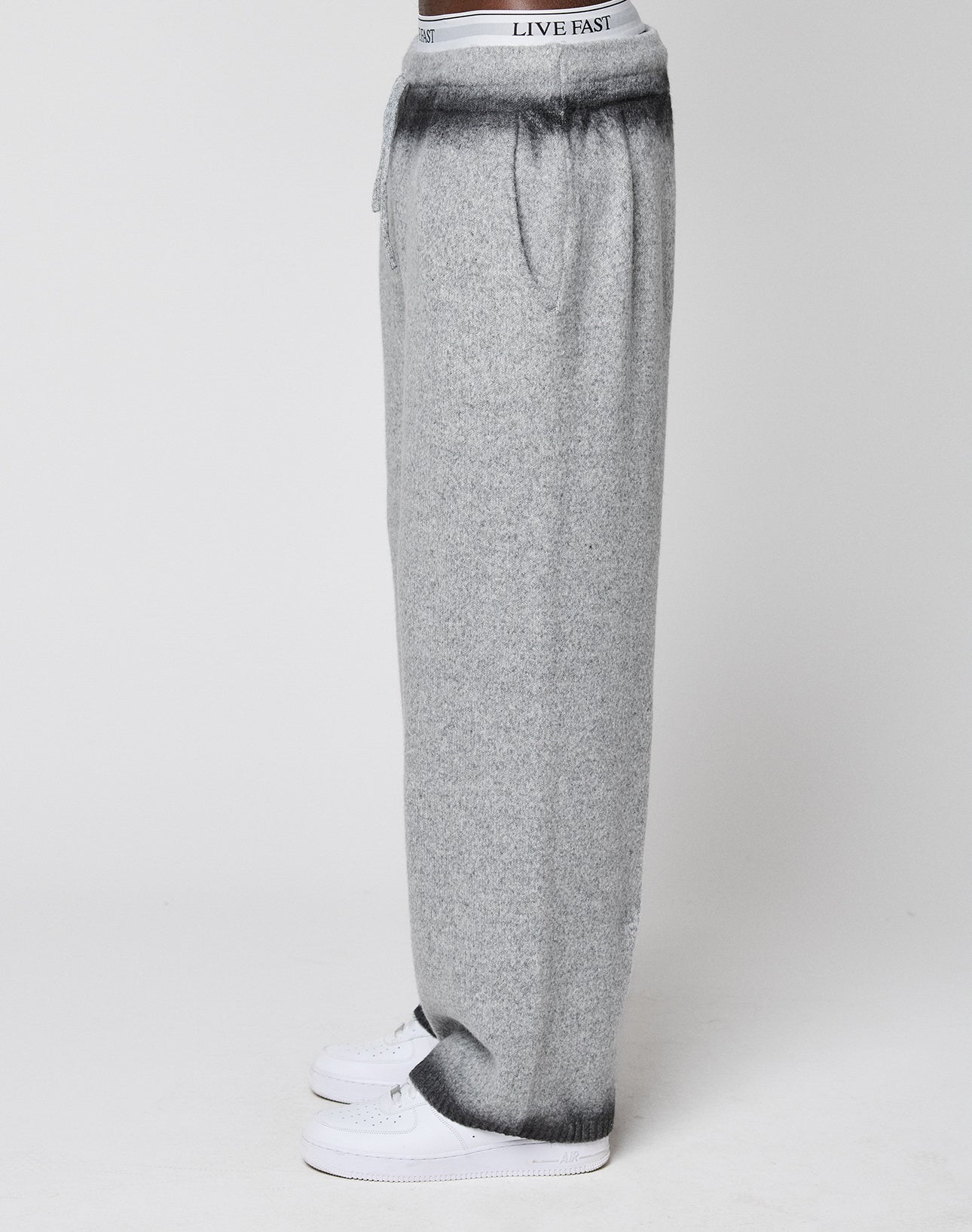 A person stands in profile wearing LIVE FAST DIE YOUNG’s Live Fast Boucle Pants—loose, wide-fit gray sweatpants with a straight leg, dark gradient waistband and cuffs, and “LIVE FAST” on the visible elastic waistband. White background.