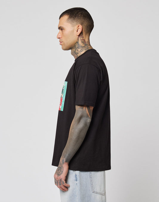 A person with short hair and tattoos stands in profile, wearing the LIVE FAST DIE YOUNG LFDY Sixers Tee—regular fit, black with a colorful front graphic—and light denim jeans against a plain white background.
