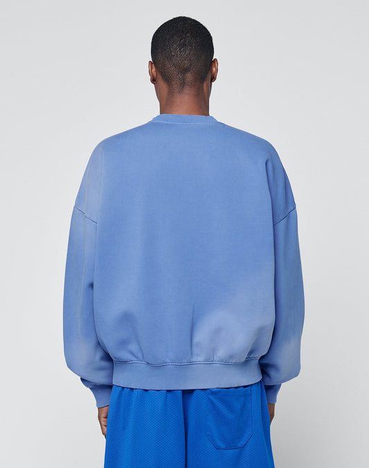 A person stands facing away from the camera, wearing a loose-fitting, light blue Basic LiveFast Sweater by LIVE FAST DIE YOUNG and blue athletic shorts against a plain light gray background.