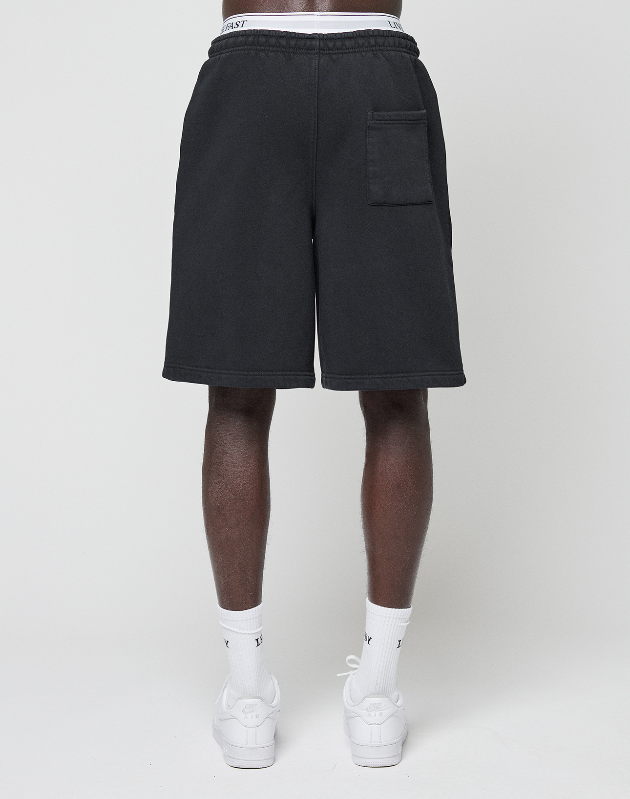 A person stands facing away from the camera wearing LIVE FAST DIE YOUNG Basic Shorts with an elastic waistband, single right back pocket, white socks, and white sneakers against a plain light background.