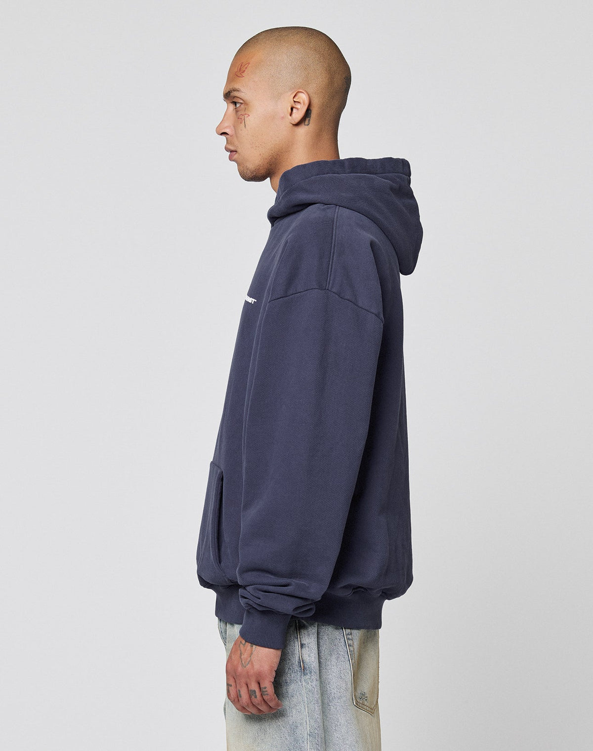A person with a shaved head wearing an oversize, dark blue LIVE FAST DIE YOUNG Live Fast Plain Hooded aus Baumwolle and light blue jeans stands in profile against a plain light gray background.