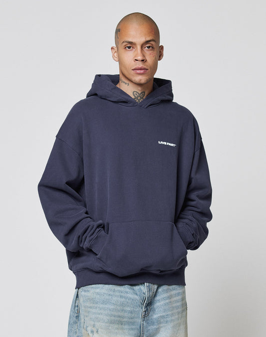 A person with a shaved head and face tattoos wears the Live Fast Plain Hooded by LIVE FAST DIE YOUNG, an oversize fit navy hoodie with "LIVIN FAST" front print, paired with light blue jeans, posing hands-in-pocket against a plain light background.