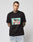 A tattooed man wears the LIVE FAST DIE YOUNG LFDY Sixers Tee, a black regular fit cotton shirt with a graphic of two cartoon people in a car and the words “LIVE FAST,” standing against a plain white background.