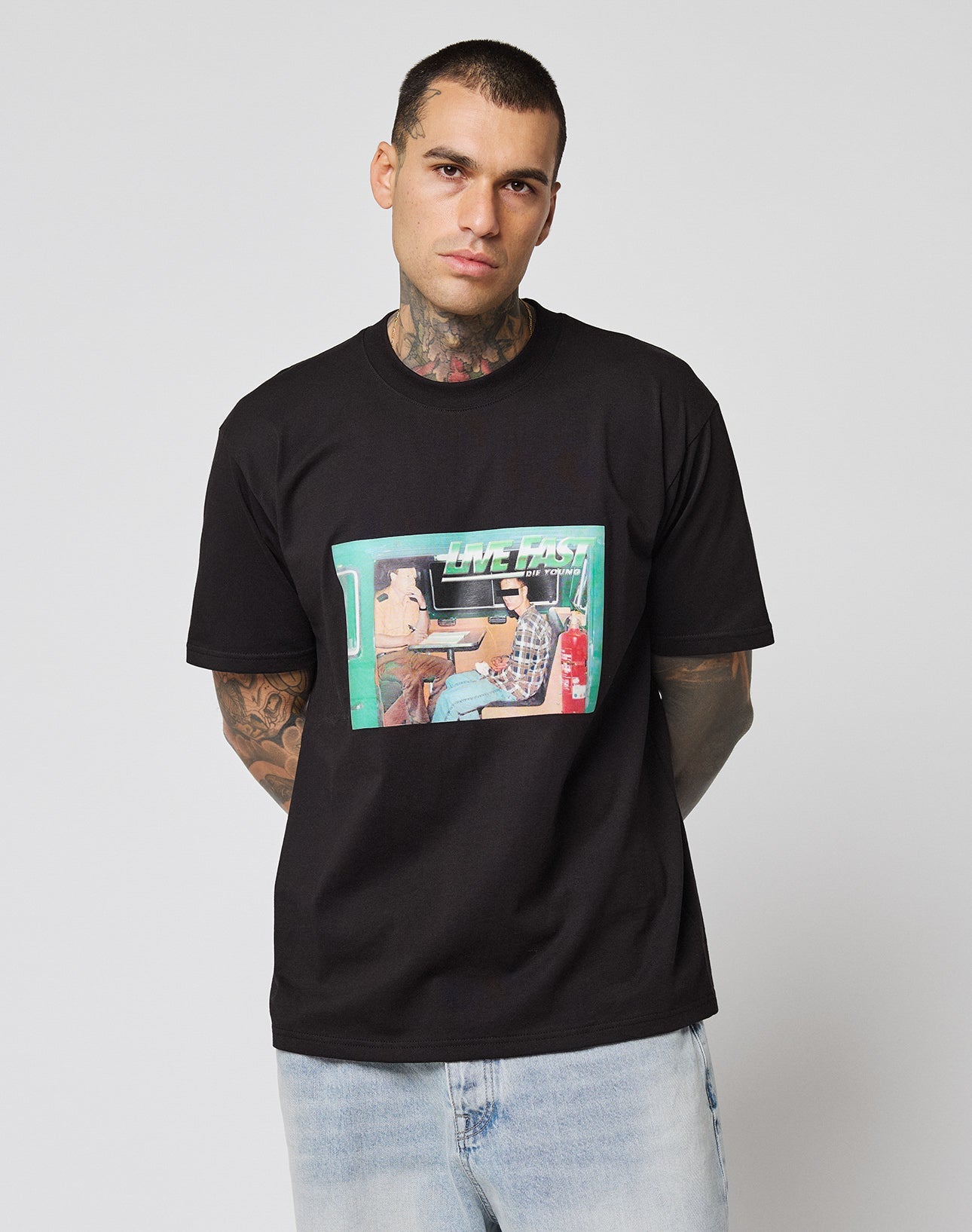 A tattooed man wears the LIVE FAST DIE YOUNG LFDY Sixers Tee, a black regular fit cotton shirt with a graphic of two cartoon people in a car and the words “LIVE FAST,” standing against a plain white background.