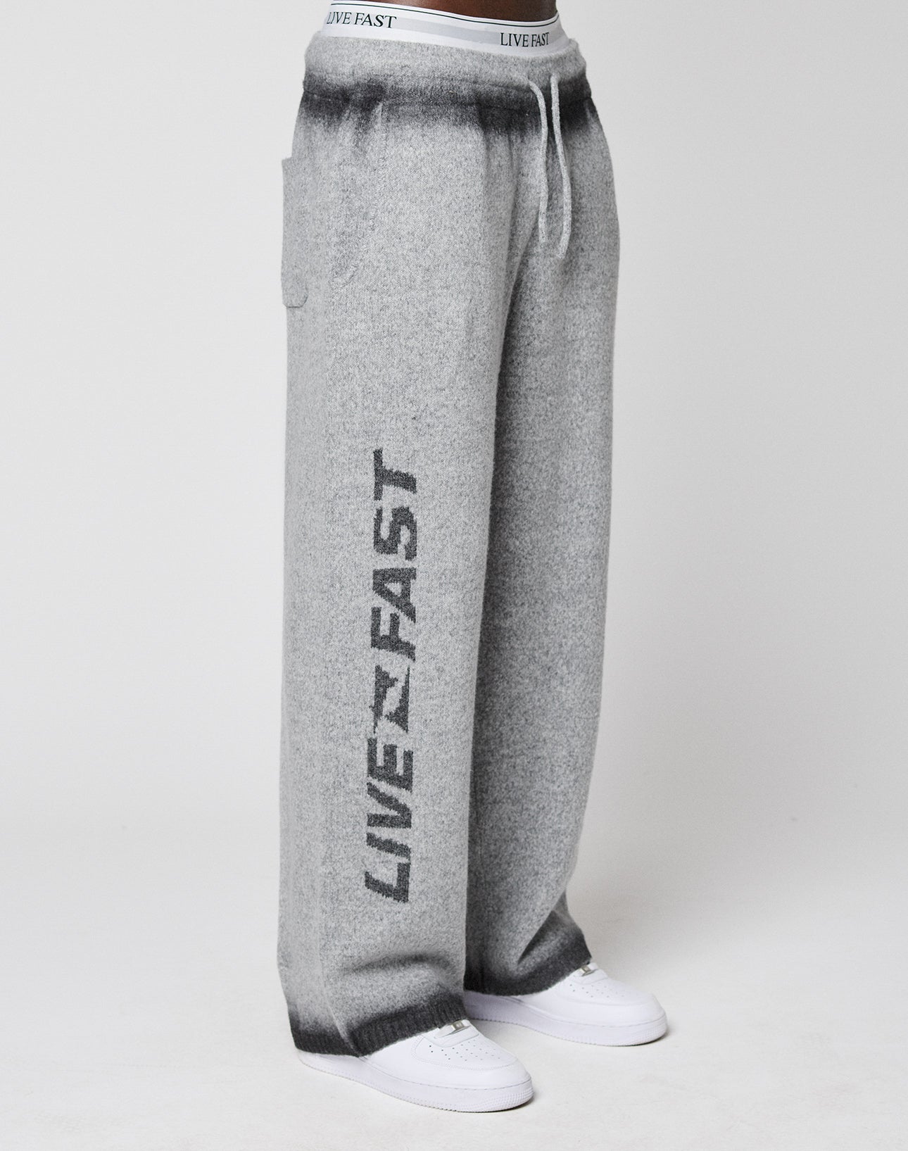 A person wears loose-fitting Live Fast Boucle Pants by LIVE FAST DIE YOUNG, in gray with "LIVE FAST" printed on the left leg and an elastic waistband. Styled with white sneakers and a straight-leg design against a plain light background.