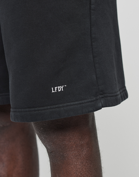 Close-up of a person wearing LIVE FAST DIE YOUNG Basic Shorts in black, featuring a regular fit and white LFDY™ logo embroidered near the hem. Only part of the shorts and the person’s upper leg are visible.