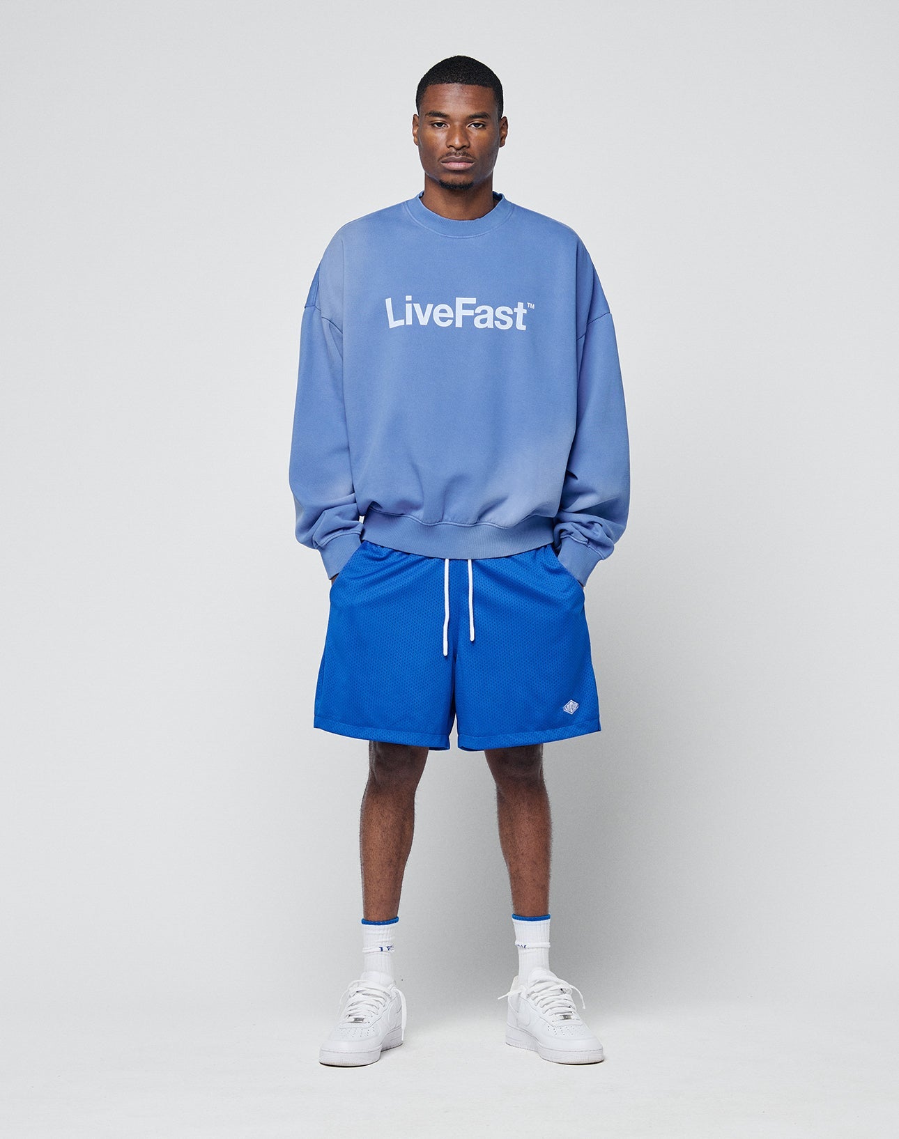 A man in a light blue Basic LiveFast Sweater by LIVE FAST DIE YOUNG, blue shorts, blue socks, and white sneakers stands against a plain light gray background with his hands in his pockets.