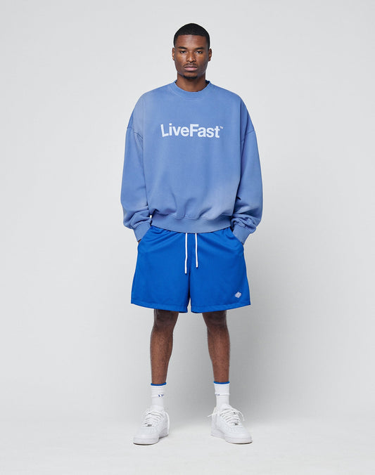 A man stands against a plain background wearing a blue LIVE FAST DIE YOUNG sweatshirt and Basic Mesh Shorts, paired with white socks and sneakers, his hands in his pockets.