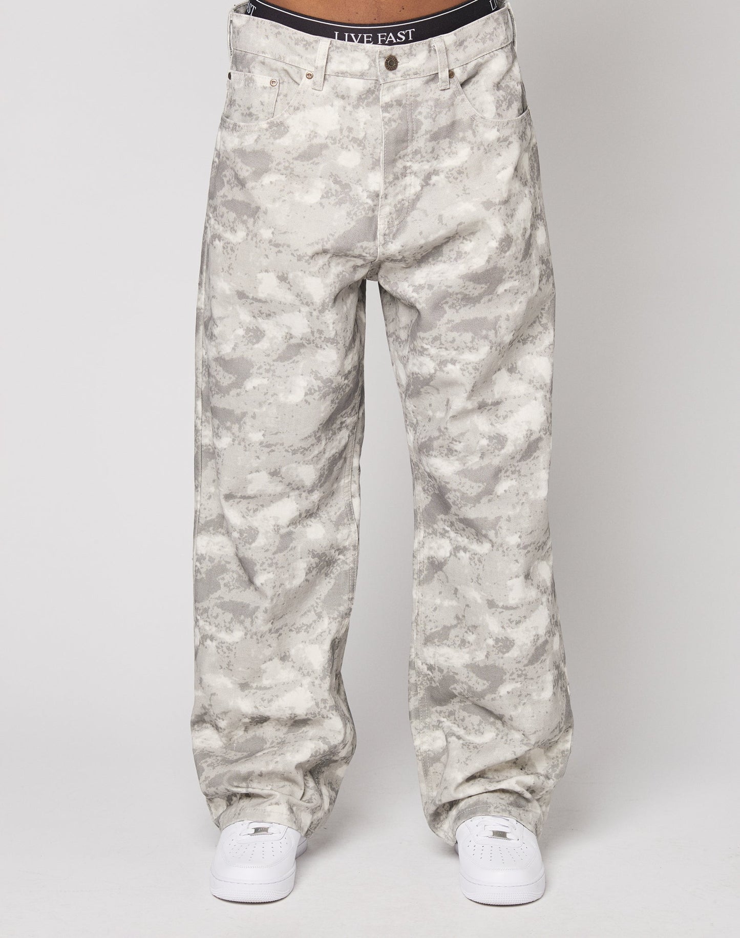A person wears LIVE FAST DIE YOUNG’s Live Fast Canvas Pants in a baggy, light gray and white camo style with a straight leg, paired with white sneakers. The waistband of black underwear featuring “LIVE FAST” text shows. Made from 100% cotton.