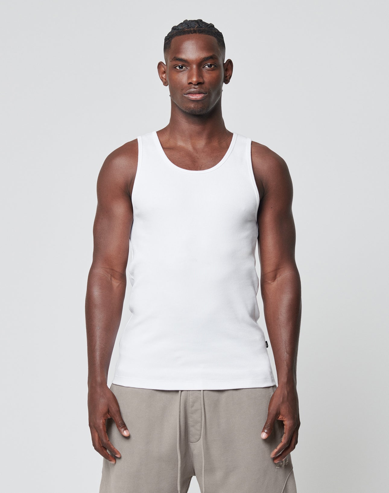 A man with a neutral expression stands against a plain background, wearing the LIVE FAST DIE YOUNG Blank Rib Tank in white and light gray shorts. His arms are relaxed at his sides.