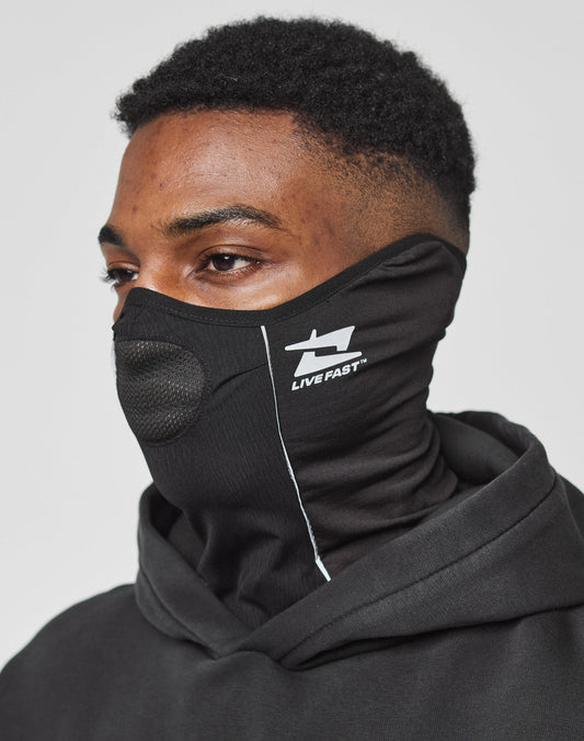 A person wears a black hooded sweatshirt and the Live Fast Performance Face Mask by LIVE FAST DIE YOUNG, featuring a mesh mouth, reflektierender Druck, and the LIVE FAST™ logo, photographed from the side against a plain background.