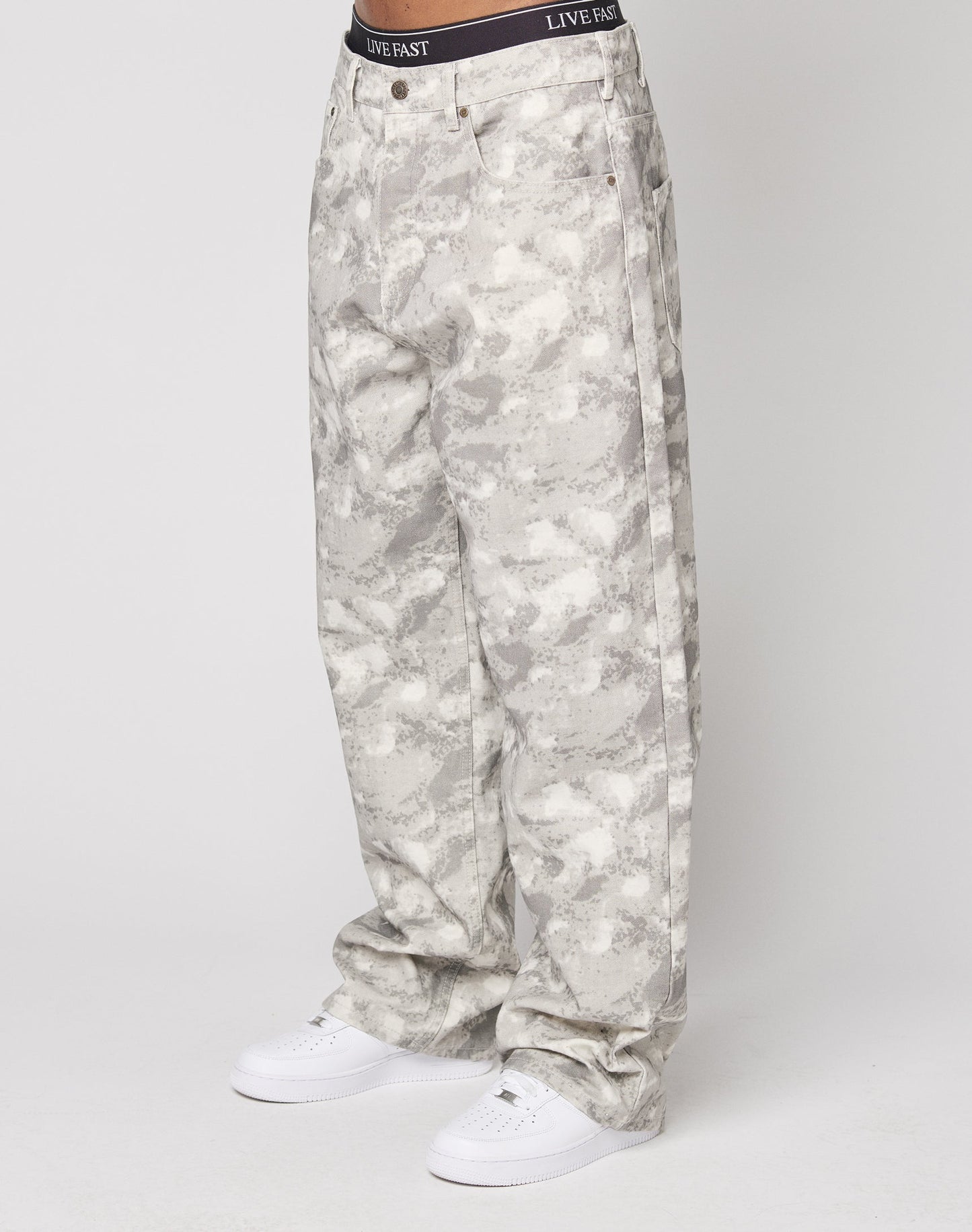 A person wears LIVE FAST DIE YOUNG’s Live Fast Canvas Pants, featuring loose-fitting light gray camouflage with a Baggy Fit and straight leg, paired with white sneakers. The waistband says LIVE FAST. The background is plain white. | model