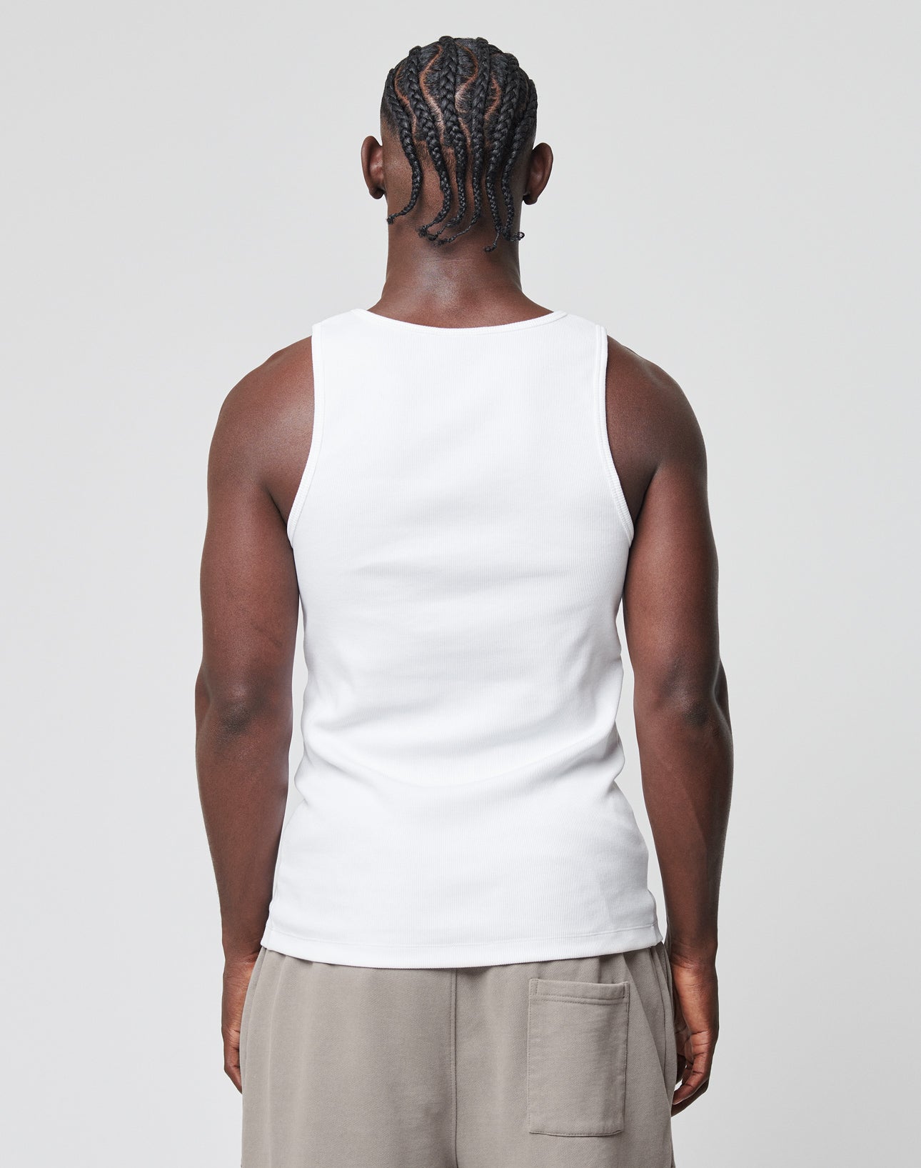 A man with braided hair is shown from the back, wearing a white LIVE FAST DIE YOUNG Basic Rib Tank and beige pocketed shorts, standing against a plain light gray background.