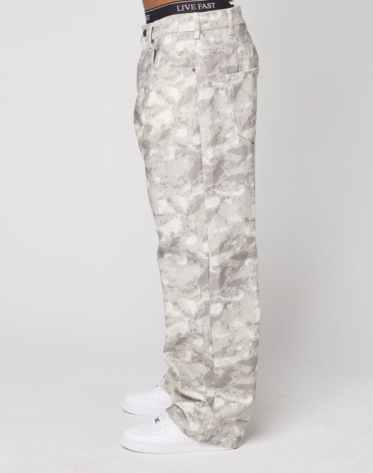 A person stands sideways in LIVE FAST DIE YOUNG’s Live Fast Canvas Pants—light gray camo with a baggy fit and wide straight leg—paired with white sneakers and a black LIVE FAST waistband, set against a plain white background.
