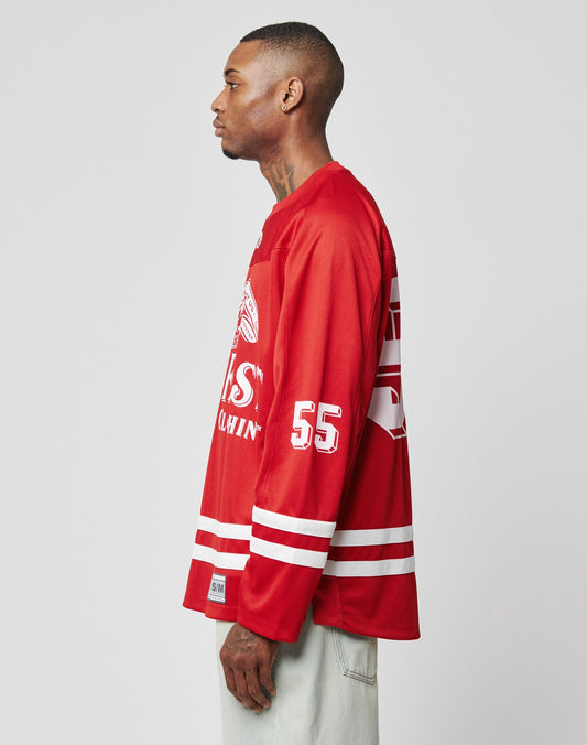 A man stands in profile against a plain gray background, wearing an oversize fit LFDY Hockey Jersey by LIVE FAST DIE YOUNG in red with white stripes and the number 55 on the sleeve, paired with pale jeans.