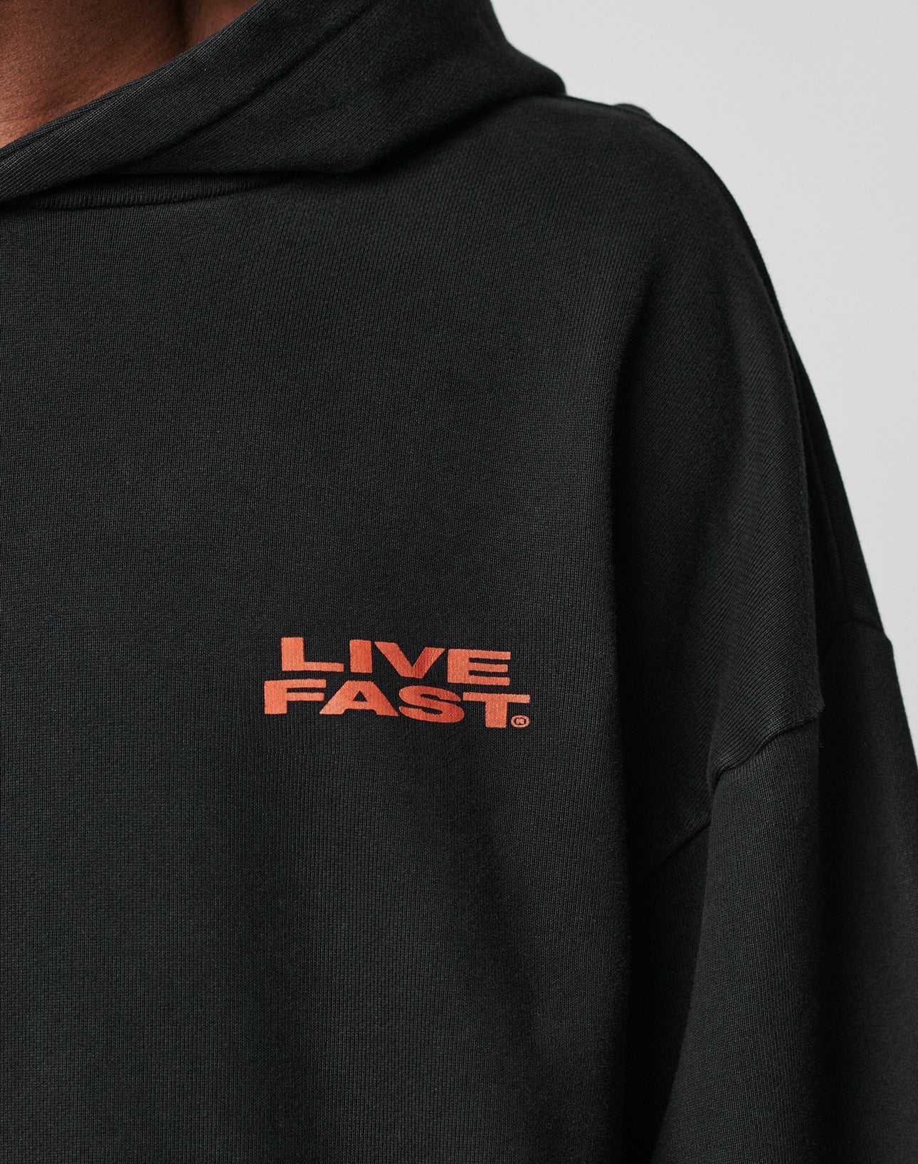 Close-up of a person in the Print Over Hooded by LIVE FAST DIE YOUNG, an oversize black hoodie made from 100% Baumwolle with LIVE FAST in bold orange on the left chest. Only part of the face and shoulder are visible against a neutral background.