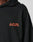 Close-up of a person in the Print Over Hooded by LIVE FAST DIE YOUNG, an oversize black hoodie made from 100% Baumwolle with LIVE FAST in bold orange on the left chest. Only part of the face and shoulder are visible against a neutral background.