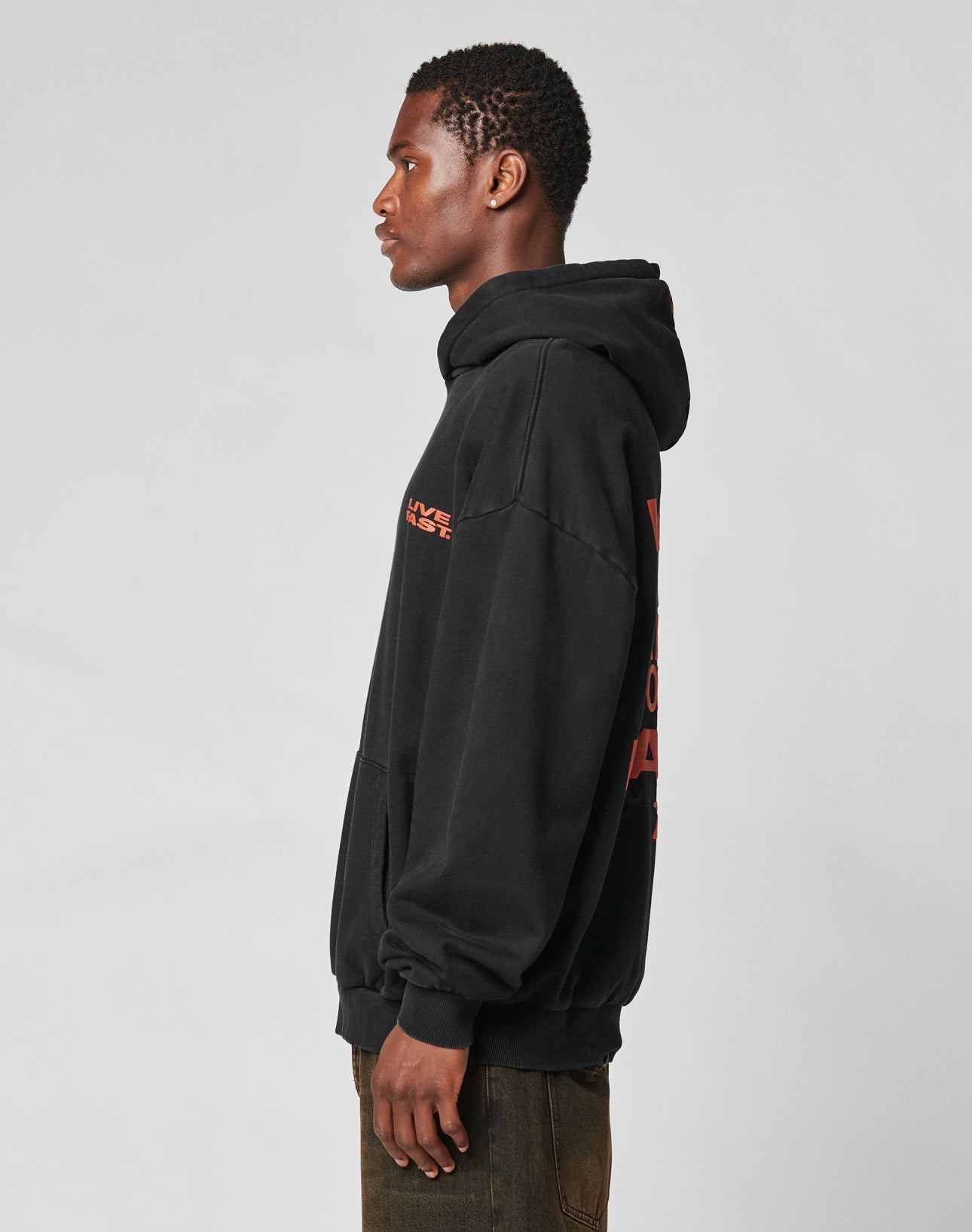 A young man in profile wears the LIVE FAST DIE YOUNG Print Over Hooded sweatshirt, oversize fit in black with red Logo-Stick on sleeve and back, paired with brown pants. Made from 100% Baumwolle for comfort and style.