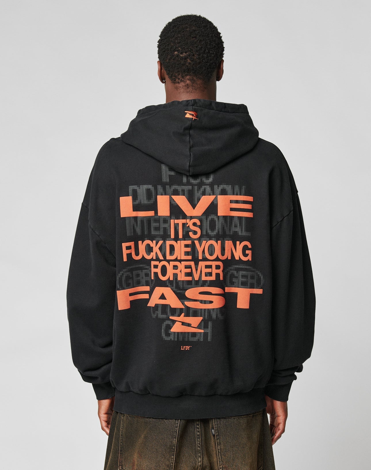 A person, facing away against a plain light background, wears an oversize fit black Print Over Hooded by LIVE FAST DIE YOUNG with bold orange LIVE ITS FUCK DIE YOUNG FOREVER FAST text on the back. Made from Baumwolle (cotton).