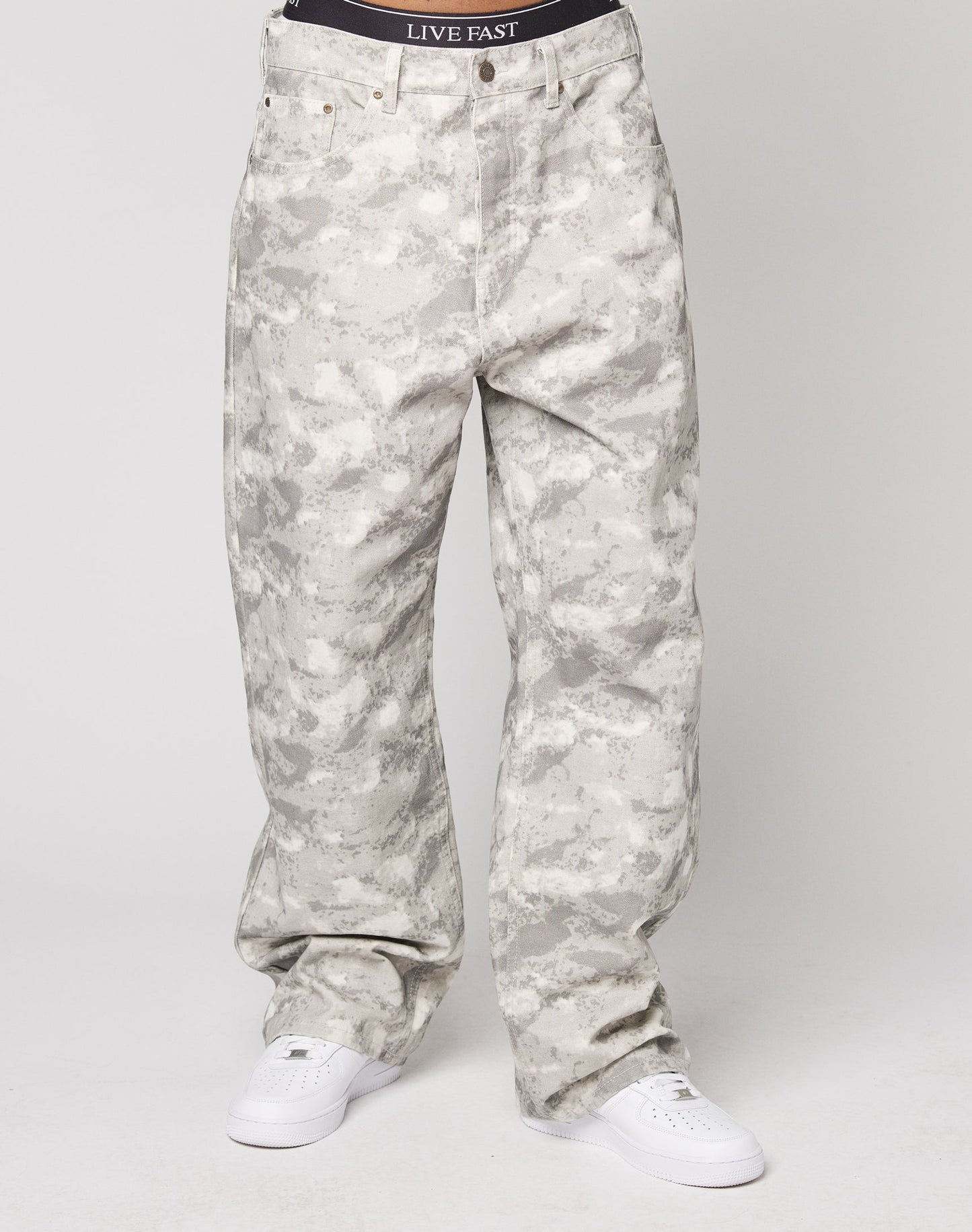A person stands against a plain white background wearing LIVE FAST DIE YOUNG’s Live Fast Canvas Pants—loose-fitting, gray and white camo, straight leg, baggy fit, 100% cotton—paired with white sneakers and black underwear with LIVE FAST on the waistband.
