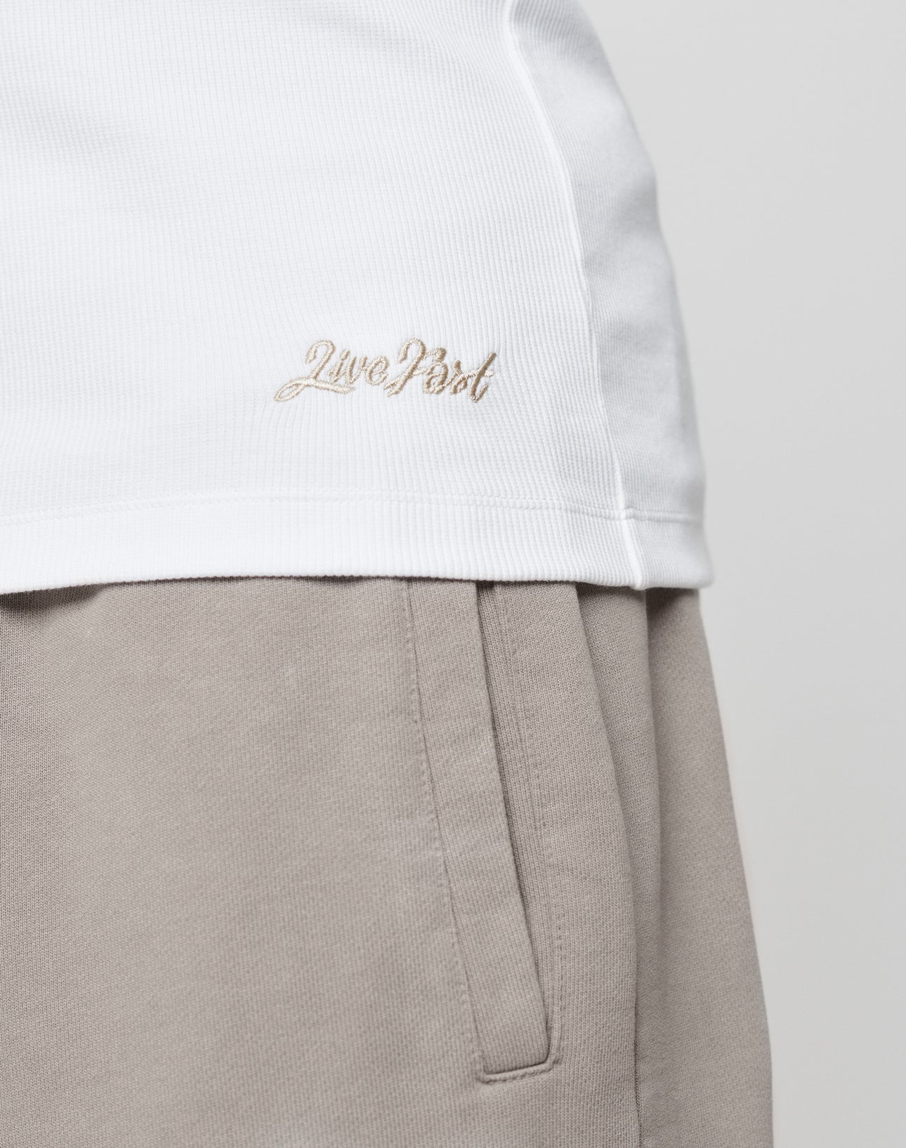 Close-up of a person wearing the LIVE FAST DIE YOUNG Basic Rib Tank in white, featuring an embroidered logo near the hem, paired with light beige pants. A plain, light background completes the look.