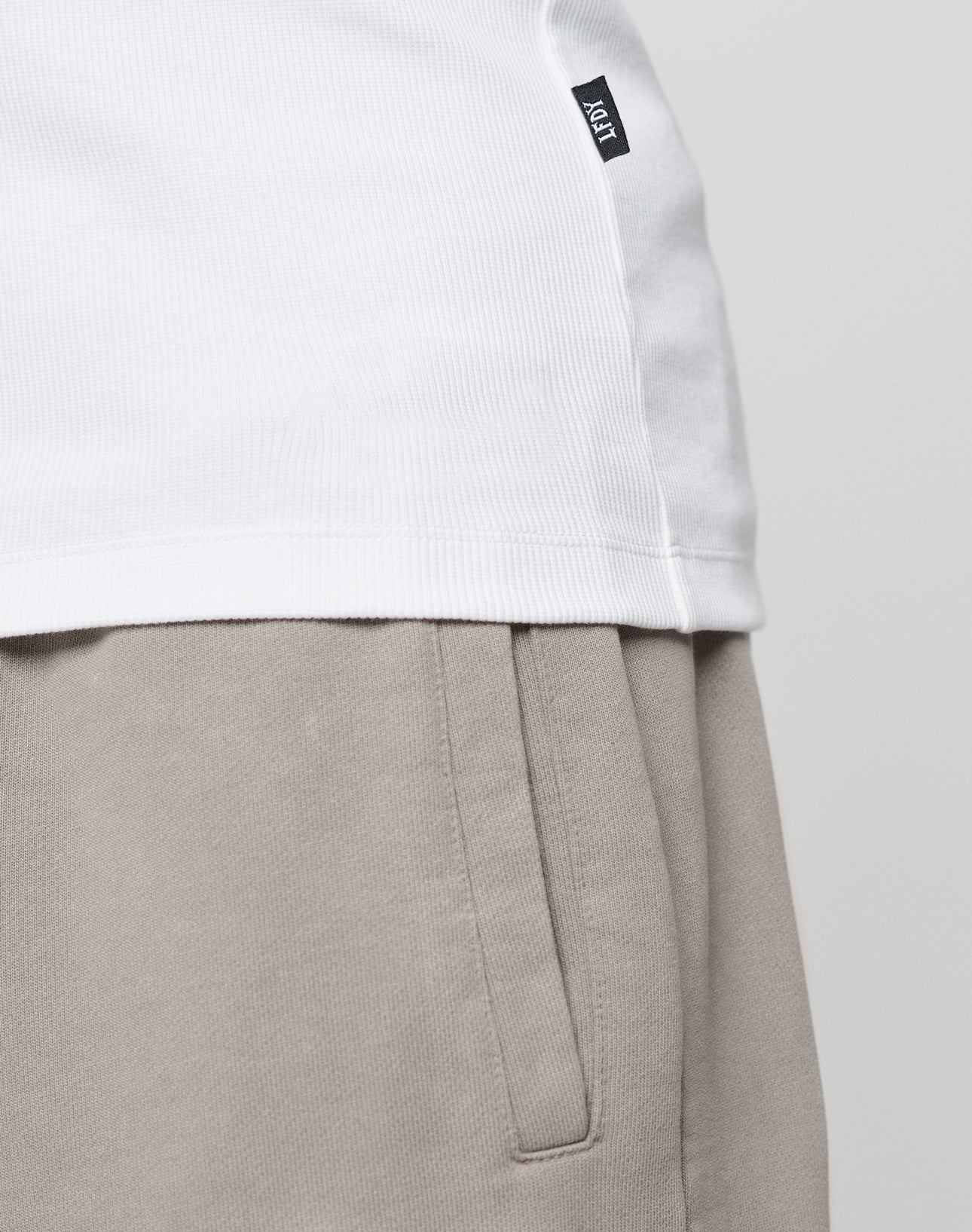 Close-up of a person wearing the LIVE FAST DIE YOUNG Blank Rib Tank in white and beige Slim Fit pants, highlighting the waistband, side pocket, and a small black tag against a plain background.