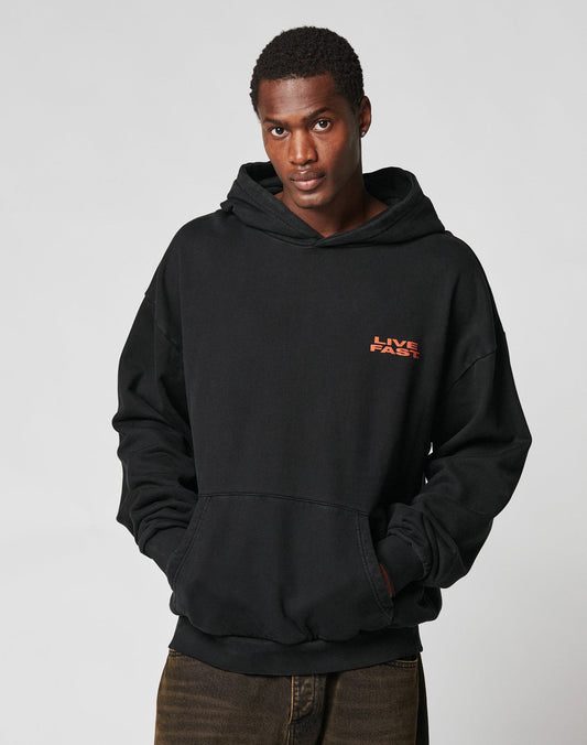 A person wears the LIVE FAST DIE YOUNG Print Over Hooded, a black oversized 100% Baumwolle hoodie with LIVE FAST in red on the chest, standing against a plain light background, hands in pockets and looking slightly upward.