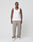 A man stands facing forward against a plain white background, wearing the LIVE FAST DIE YOUNG Basic Rib Tank in slim fit white, paired with loose beige pants and white sneakers. His arms are relaxed by his sides.