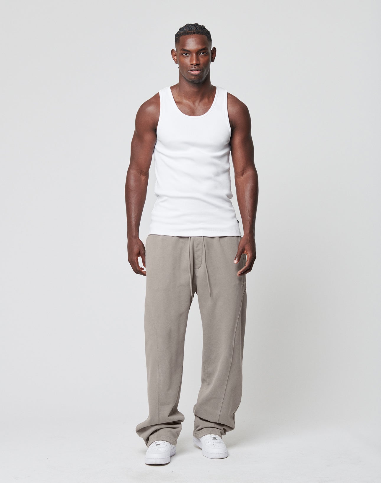 A man faces forward against a plain background, wearing the LIVE FAST DIE YOUNG Blank Rib Tank in white, paired with loose beige pants and white sneakers. His expression is neutral, with arms resting at his sides.