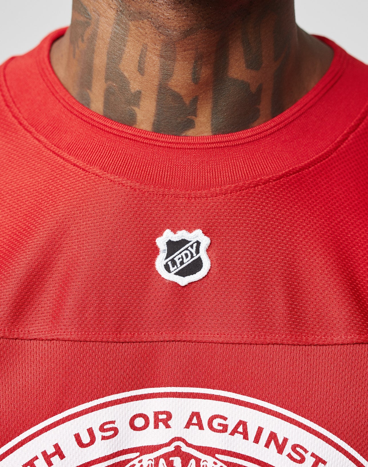 A close-up of a person in a red LFDY Hockey Jersey by LIVE FAST DIE YOUNG, featuring an oversize fit, black chest badge, bold neck tattoos, and an eye-catching Ärmelprint.