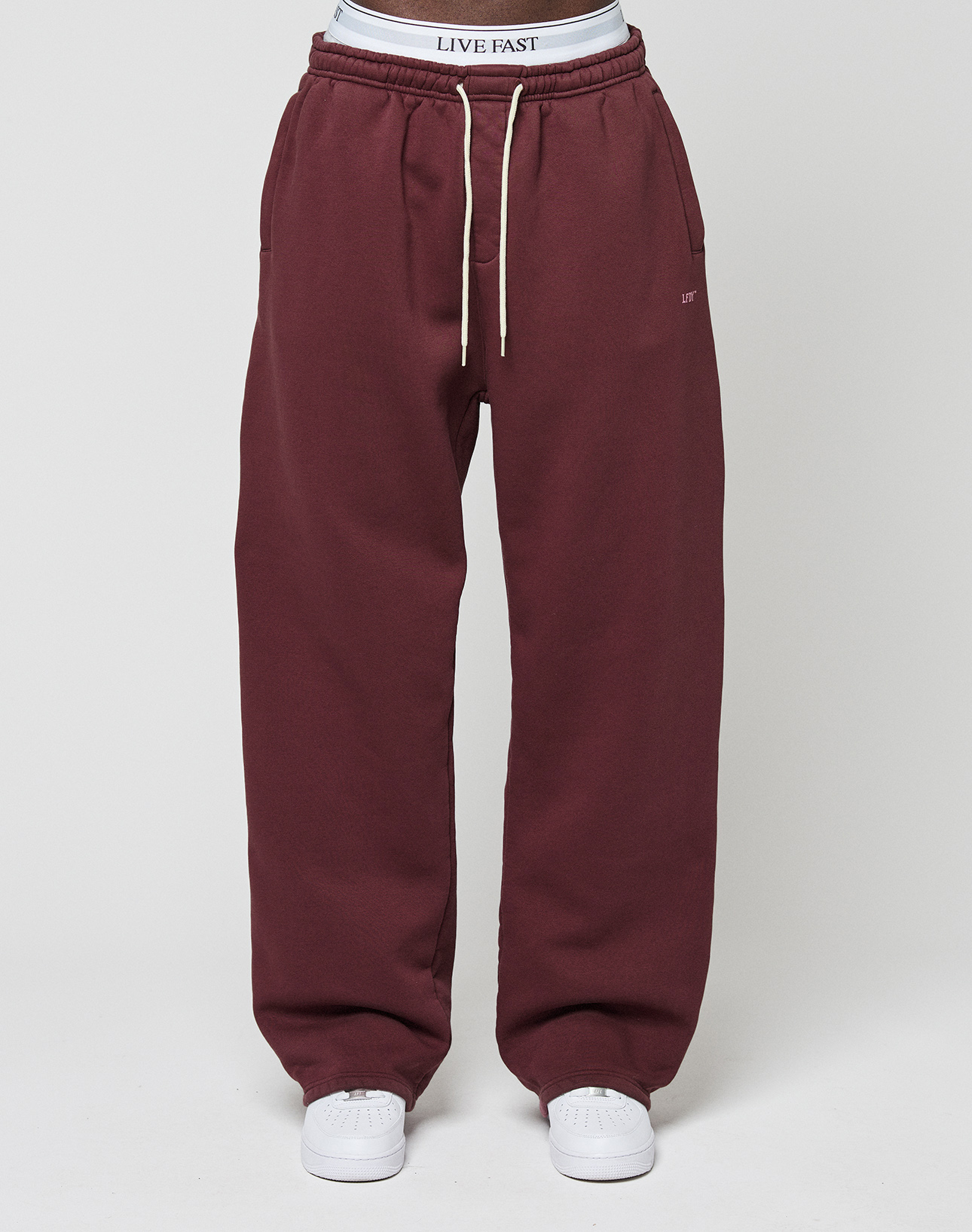 Model in maroon LIVE FAST DIE YOUNG Basic Wide Jogger with white drawstrings, logo detailing, side pockets, paired with white sneakers and a visible LIVE FAST waistband, poses against a plain light background.