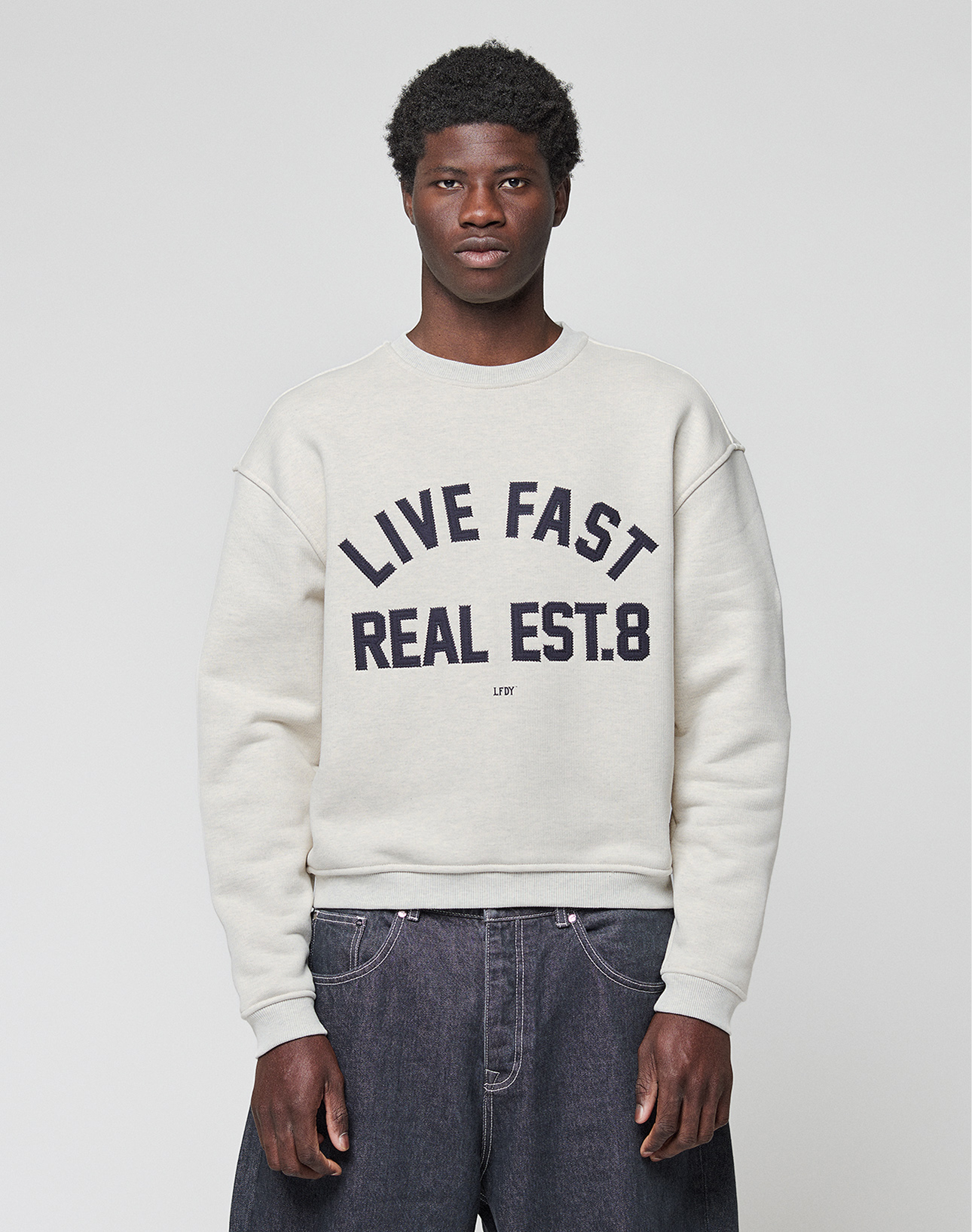 A person stands against a plain background wearing the LFDY Heavyweight Lounge Sweater by LIVE FAST DIE YOUNG, featuring an oversized 100% Baumwolle fit and bold "LIVE FAST REAL EST.8" text on the front, paired with loose dark jeans. | model