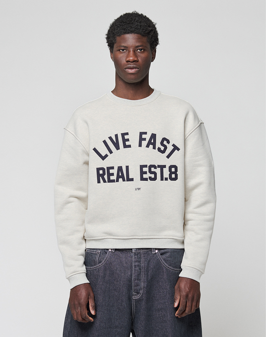 A person stands against a plain background wearing the LFDY Heavyweight Lounge Sweater by LIVE FAST DIE YOUNG, featuring an oversized 100% Baumwolle fit and bold "LIVE FAST REAL EST.8" text on the front, paired with loose dark jeans. | model